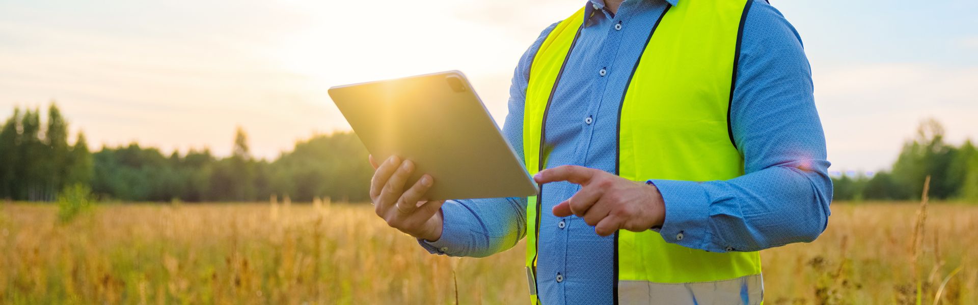 Henkilö, jolla on huomioliivi päällään pitää kädessään tablet-tietokonetta. Taustalla paistaa aurinko ja on peltomaisema.